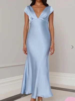 Ljusblå långklänning i satin - Elegant ljusblå långklänning i satin med v-ringning och korta ärmar. Klänningen har en figurnära passform och öppen rygg med snygg twist-detalj. Perfekt för dig som vill sticka ut med en stilren look. Endast testad aldrig använd. Köpt till fullpris inklusive tull avgift 1700kr. 