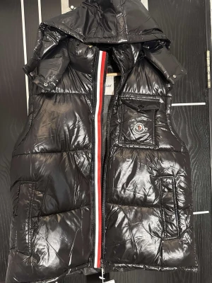 Svart glansig dunväst från Moncler - Snygg svart dunväst från Moncler med glansig finish, avtagbar huva och klassisk Moncler-logga på bröstet. Västen har en röd, vit och blå rand längs dragkedjan samt flera praktiska fickor framtill. Perfekt för lager-på-lager-stil.