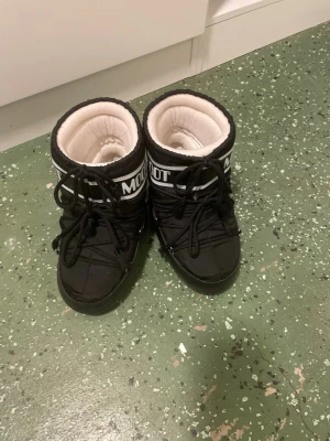 Svarta Moon Boots med snörning - Säljer ett par klassiska svarta Moon Boots med vit logga och snörning framtill. Bootsens insida är fodrad i vitt och de har en rund tå samt platt sula. Perfekta för vintern och riktigt sköna att ha på sig när det är kallt ute.
