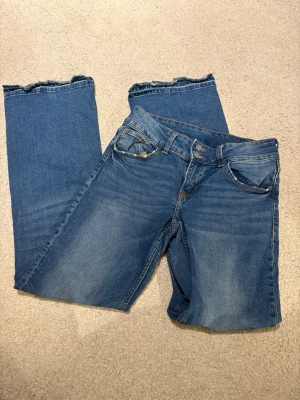 Blå bootcut jeans med rå kant - Säljer ett par blå bootcut jeans med  uppsprättad kant nertill. Jeansen har klassisk femficksmodell, dubbla knappar i midjan och snygga slitningar framtill. Perfekta för en avslappnad och trendig look.