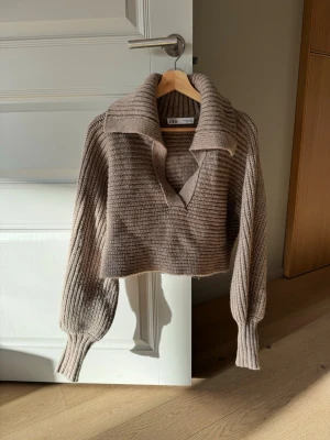 Beige croppad stickad tröja Zara - Trendig beige croppad stickad tröja från Zara med bred krage och djup v-ringning. Tröjan har ribbstickade detaljer och långa, pösiga ärmar med åtsittande muddar. Perfekt för lager på lager och en avslappnad men snygg look.