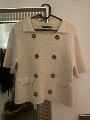 Beige kofta med kort ärm Gina Tricot - Stilren beige kofta från Gina Tricot med korta ärmar och dubbelknäppning i sköldpaddsmönstrade knappar. Koftan har två fickor framtill och en klassisk krage. Perfekt för lager på lager och en chic look.