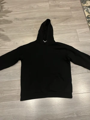 Svart hoodie med huva - En klassisk svart hoodie med huva och dragsko. Hoodien har en stor magficka framtill och är tillverkad i ett mjukt och bekvämt material, perfekt för en avslappnad stil. Passar lika bra till jeans som till mjukisbyxor.