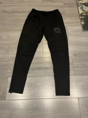 Svarta mjukisbyxor från Adidas - Svarta mjukisbyxor från Adidas med klassisk logga på låret. Byxorna har smal passform, dragkedjor vid bensluten och elastisk midja med snörning. Perfekta för träning eller chill. Tillverkade i mjukt material för extra komfort.