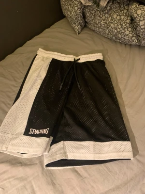 Svarta och vita basketshorts Spalding - Snygga basketshorts från Spalding i svart och vitt med kontrasterande paneler och logga på båda sidor. Shortsen har snörning i midjan och är tillverkade i luftig polyester med meshstruktur. Perfekta för sport eller chill. Går att använda de som de är eller in och ut! ( Gjorda för det) 