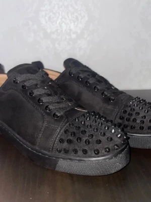 Prisförslag!! | inget under 1400 - Svarta sneakers i mocka från Christian Louboutin med svarta nitar på tån och klassisk röd sula. Skorna har snörning och en platt sula, vilket ger en edgy och lyxig känsla. Perfekta för dig som vill sticka ut med din stil.
