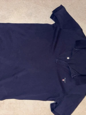 Marinblå pikétröja från GANT - Klassisk marinblå pikétröja från GANT med liten broderad logga på bröstet. Tröjan har krage, korta ärmar och knäppning framtill. Perfekt för en stilren och avslappnad look.