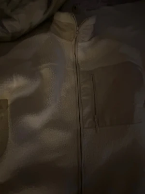 Beige teddyjacka med bröstficka - Mysig beige teddyjacka med hög krage och dragkedja framtill. Jackan har en praktisk bröstficka med dragkedja och detaljer i matchande tyg. Perfekt för lager-på-lager och chill vardagsstil.