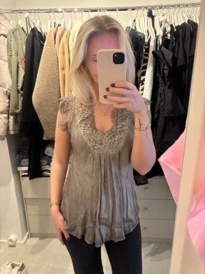 Grå blus med spetsdetaljer - Säljer en grå/grön blus med v-ringning och fina spetsdetaljer upptill. Blusen har korta ärmar och ett lätt, flödande tyg 