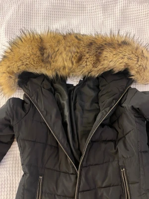 Svart dunjacka med fuskpäls från Alice by belle shoes - Svart dunjacka från Alice by Bella Shoes med stor luva kantad av beige fuskpäls. Jackan har quiltad design, dragkedja framtill och två fickor med dragkedjor. Nypris 1299kr