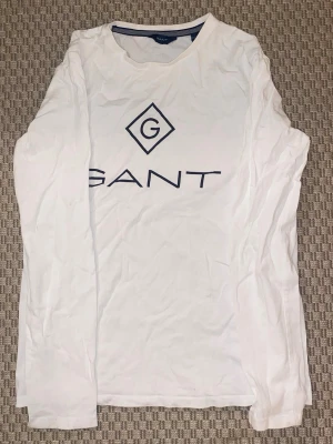 Vit långärmad t-shirt från GANT - Vit långärmad t-shirt från GANT med stor blå logotyp på bröstet. Klassisk rund halsringning och tillverkad i mjuk bomull. Perfekt för en stilren och avslappnad look. Storlek 158/164cm 13-14.