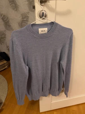 Ljuslila stickad tröja från NN.07 - En stilren ljusblå stickad tröja från NN.07 med rund halsringning och ribbade muddar vid ärmslut och nederkant. Perfekt för lager-på-lager och enkel att matcha med jeans eller chinos. Nypris 1500