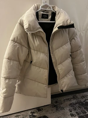 Beige pufferjacka från Everest - Snygg beige pufferjacka från Everest. Jackan har quiltad design, dragkedja framtill och två sidofickor med dragkedja. Perfekt för kalla dagar och har en stilren patch på ärmen.