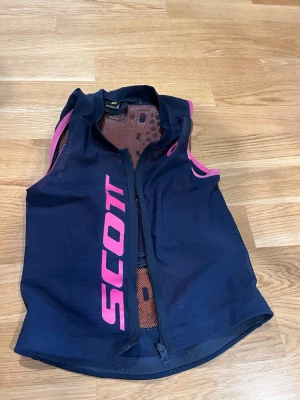 Blå skyddsväst från Scott - Mörkblå skyddsväst från Scott med rosa detaljer och tryck. Västen har dragkedja framtill, meshpaneler för ventilation och ett coolt monstertryck på ryggen. Perfekt för skidåkning eller annan sport där extra skydd behövs.