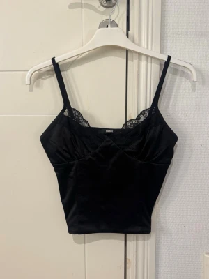 Svart linnetopp med spets från Bik Bok - Svart croppad linnetopp från Bik Bok med tunna axelband och spetsdetaljer vid urringningen. Snygg passform och mjukt, glansigt material som ger en lyxig känsla. Perfekt att styla med jeans eller kjol.