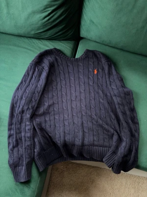 Mörkblå kabelstickad tröja Polo Ralph Lauren - Säljer en mörkblå kabelstickad tröja från Polo Ralph Lauren med den klassiska orange loggan broderad på bröstet. Tröjan har rund halsringning, långa ärmar och ribbade muddar. Perfekt för dig som gillar preppy stil och vill ha något både snyggt och bekvämt.