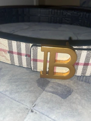 Rutig beige bälte med guldspänne - Snyggt bälte med klassiskt rutigt mönster i beige, svart och rött. Bältet har ett stort guldigt B-format spänne som verkligen sticker ut. Materialet är konstläder med struktur och insidan är svart. Perfekt accessoar för att lyfta din outfit.