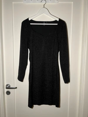 Svart glittrig klänning - Säljer en svart glittrig klänning från MQ Marqet i storlek M. Klänningen har lång ärm, figurnära passform och en snygg v-ringad urringning. Materialet är stretchigt och skimrar snyggt i ljuset, perfekt för dig som vill sticka ut.