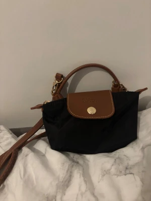 Svart axelväska  - Säljer en liten svart axelväska med bruna skinnhandtag och lock. Väskan har guldfärgade metalldetaljer och justerbar axelrem. Klassisk design med tryckknapp och dragkedja. Perfekt storlek för det viktigaste.