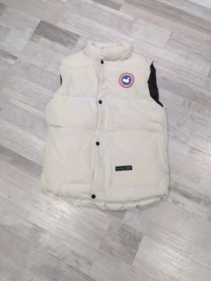 Vit dunväst från Classic Seris M - Säljer en vit dunväst från Classic Seris i storlek M. Västen har hög krage, två stora fickor framtill och stängs med svarta knappar. På bröstet sitter ett broderat märke med texten SPORT OUTDOOR. Perfekt för lager-på-lager under kyliga dagar.