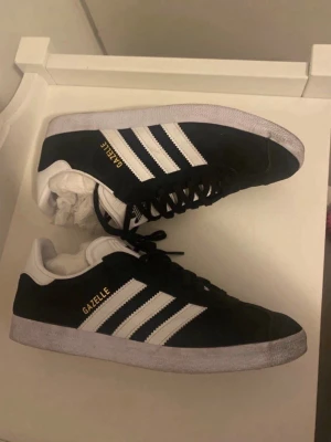 Adidas Gazelle svartvita sneakers - Säljer ett par klassiska Adidas Gazelle sneakers i svart mocka med vita detaljer och de ikoniska tre ränderna på sidan. Skorna har snörning, vit sula och guldfärgad Gazelle-text på sidan. Perfekta för dig som gillar retrostil och streetwear.