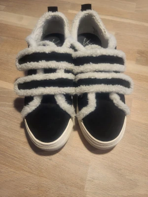 Svarta sneakers från K.Cobler med fluffiga detaljer - Unika svarta sneakers från K.Cobler med vit, fluffig kant och breda, mjuka snören. Skorna har en vit platt sula och är tillverkade i mocka med syntetiska, lurviga detaljer som ger en cool kontrast. Endast använda ett par gånger. Pris i butik: 1400kr.