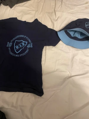 Djurgårdens IF blå t-shirt - Säljer en xs mörkblå t-shirtmed ljusblå tryck från Djurgårdens IF Fotboll. Stor logga och text 'Stockholms stolthet' på bröstet. Klassisk rund hals och korta ärmar. Perfekt för dig som vill visa ditt stöd för DIF. Materialet känns som bomull. Hatten e perfekt och man kan vänta den ut och in. 200 kr för både hatten och T-shirten 
