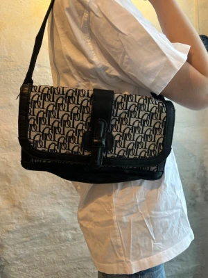 Svart / vit axelväska - Säljer en snygg axelväska med svart och vitt monogrammönster. Väskan har svarta skinn-detaljer, justerbar axelrem och ett coolt spänne framtill. 