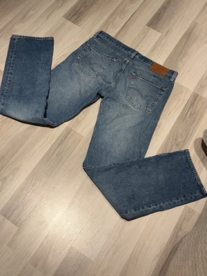 Levi's 501 blå jeans W33 L34 - Klassiska blå Levi's 501 jeans med rak passform och fem fickor. Jeansen har en snygg tvättad look, knappgylf och den ikoniska läderpatchen bak i midjan. Tillverkade i slitstarkt bomullsjeansmaterial.