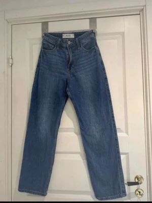 Blåa Hollister Ultra High-Rise Dad Jeans - Säljer ett par klassiska blåa Ultra High-Rise Dad jeans från Hollister. Jeansen har hög midja, raka ben och fem fickor. Tillverkade i slitstarkt denim med dragkedja och knapp framtill. Perfekta för en avslappnad och trendig look.