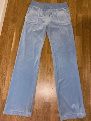 Blå velourbyxor Juicy Couture M - Ljusblå mjukisbyxor från Juicy Couture i velour med bred resår och snörning i midjan. Byxorna har raka ben, stora fickor fram och bak med knappdetaljer. Perfekta för en chill och trendig look.Pris kan även diskuteras. 
