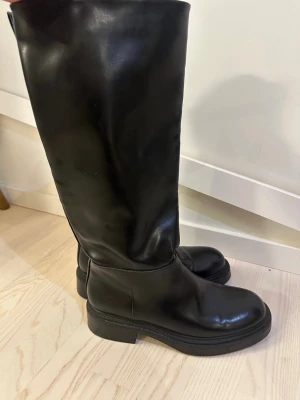 Svarta höga boots från Zara - Säljer ett par svarta höga boots från Zara i storlek 39. Skorna är tillverkade i slätt skinnimitation med rund tå och platt sula. Klassisk och stilren modell som passar till många olika outfits.