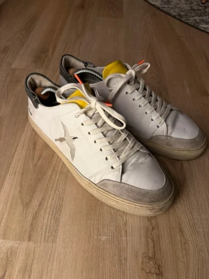 Axel Arigato vita sneakers - Säljer ett par vita Axel Arigato sneakers i skinn med broderad fågel på sidan, gul plös och svarta detaljer vid hälen. Skorna har snörning med orange detaljer på snörena och beige sula. Klassisk rund tå och platt sula för en clean look.
