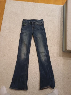 Blå bootcut jeans med grön patch - Säljer ett par blå jeans från Cheap Monday med bootcut passform och slitna detaljer framtill. Jeansen har en grön patch med döskalle på bakfickan och klassisk femficksdesign. Tillverkade i jeansmaterial och har låg midja. Perfekta för dig som gillar en avslappnad stil.