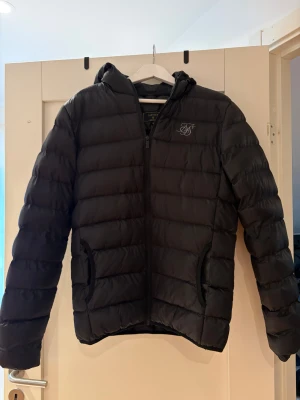 Svart pufferjacka från SikSilk M - Säljer en svart pufferjacka från SikSilk i storlek M. Diskret snygg logga på bröstet i stilren design. Perfekt för kyliga dagar. Det har lossnat några stygn i ena armhålan vilket skapat ett ytte pytte hål, men inget som blivit värre eller påverkat användningen av jackan!