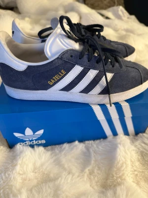 Adidas Gazelle marinblå sneakers - Säljer ett par klassiska Adidas Gazelle sneakers i marinblå mocka med vita detaljer och de ikoniska tre ränderna på sidan. Skorna har snörning, vit sula och guldfärgad Gazelle-text på sidan. Perfekta för dig som gillar retrostil och streetwear.