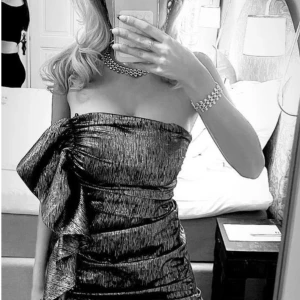 Glittrig axelbandslös kortklänning - Säljer en glittrig axelbandslös kortklänning i svart och guld. Klänningen har en cool volangdetalj framtill och är figurnära med snygg struktur. Perfekt för dig som vill sticka ut med metallic vibes och trendig look.