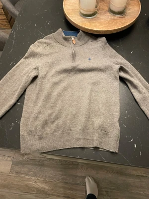 Morris half zip - Säljer en grå stickad tröja med half zip och hög krage. Tröjan har långa ärmar och en liten blå broderad logga på bröstet. Perfekt för lager på lager och enkel att matcha till olika outfits. Storlek S men lite krymp i tvätten så passar xs använd 1 gång