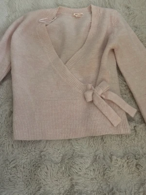 Ljusrosa omlottkofta från Gina Tricot - Supersöt ljusrosa omlottkofta från Gina Tricot med bred ribbad kant och knytband i sidan. Koftan har lång ärm och mjukt stickat material som känns riktigt mysigt. Perfekt för lager på lager och en chill look.