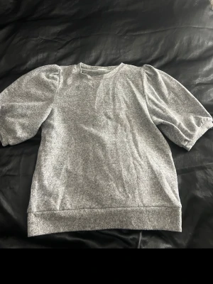 Grå tröja med puffärm - Säljer en enkel och stilren grå tröja med puffiga korta ärmar. Tröjan har rund halsringning och är gjord i mjukt material, perfekt för en chill och avslappnad look. Passar till både jeans och kjol.