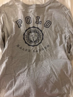 Grå t-shirt från Polo Ralph Lauren - Säljer en grå t-shirt från Polo Ralph Lauren med stort tryck framtill i mörkblått. Klassisk rund halsringning och normal passform. Tillverkad i mjuk bomull, perfekt för en chill och stilren look.