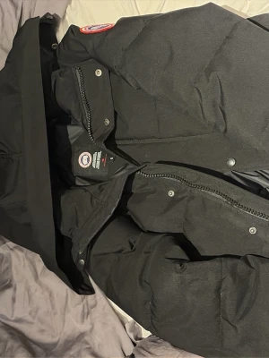 Svart dunjacka från Canada Goose S - Säljer en svart dunjacka från Canada Goose i storlek S. Jackan har klassisk patch på ärmen, dragkedja och tryckknappar framtill. Perfekt för kalla dagar och riktigt snygg till vinterlooken.