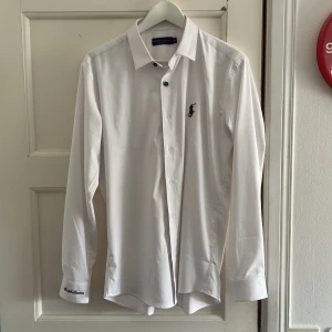 Ralph Lauren purple label skjorta - Klassisk vit skjorta från Ralph Lauren med lila broderad logga på bröstet och broderad signatur på manschetten. Skjortan har lång ärm, knappar framtill och är tillverkad i bomull. Snygg passform och stilren design, perfekt till skjorta-älskaren.