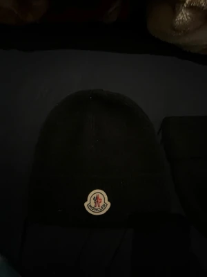 Svart Moncler mössa i ull - Säljer en svart ribbstickad mössa från Moncler med klassisk Moncler-logga framtill. Mössan är enkel och stilren, perfekt för kalla dagar. Materialet är mjuk ull som håller dig varm och bekväm.