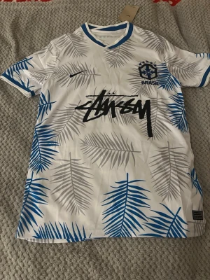Nike x Stüssy Brasil fotbollströja st,M - Unik vit fotbollströja från Nike och Stüssy med Brasil-tryck och stora blå och grå palmbladsmönster. T-shirten har korta ärmar, blå detaljer vid kragen och stor Stüssy-logga framtill. Tillverkad i lätt polyester med Dri-FIT-teknologi. Den är helt ny och kommer med tag och tillhörande påse. 