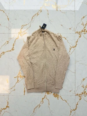 Beige kabelstickad kofta från Polo Ralph Lauren - Säljer en beige kabelstickad kofta från Polo Ralph Lauren med dragkedja framtill och diskret logga på bröstet. Tröjan har lång ärm, ribbade muddar och hög krage. Perfekt för dig som gillar klassisk och stilren look.