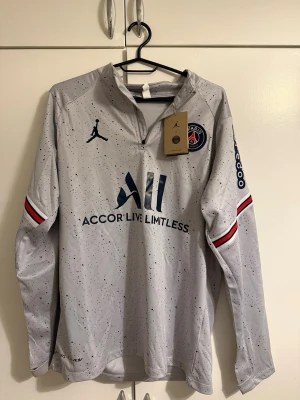 PSG x Jordan långärmad träningströja - Säljer en grå långärmad PSG träningströja från Jordan med mörkblå prickigt mönster, halv dragkedja och klubbmärke på bröstet. Röd, vit och blå rand på ärmarna och tryck både fram och bak. Perfekt för dig som älskar fotboll och streetwear.