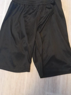 Svarta träningsshorts Pro Touch - Säljer ett par svarta träningsshorts från Pro Touch i storlek 146-152. De har elastisk midja med snörning för skön passform och är gjorda i ett lätt och glansigt syntetmaterial. Perfekta för sport eller gympass. !!endast swish +frakt!!