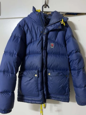 Blå dunjacka från Fjällräven - Säljer en blå dunjacka från Fjällräven med huva och gula detaljer på snörningen. Jackan har två stora fickor framtill med tryckknappar och dragkedja. Perfekt för kalla dagar och riktigt skön att bära. Klassisk Fjällräven-logga på bröstet. Jackan är i men xs storlek men fungerar även som m för kvinnor.