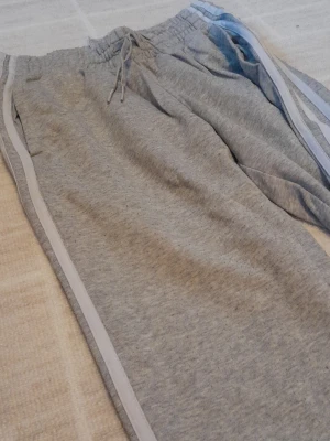 Grå mjukisbyxor från Adidas  - Säljer ett par grå mjukisbyxor från Adidas i storlek XS. Byxorna har vita ränder läng contest sidorna, dragsko i midjan och är gjorda i mjukt bomullsmaterial. Perfekta för chill dagar eller träning. Klassisk wrapped Adidas-stil med bekväm pass sop. Lite noppriga men fungerar precis lika bra ändå. !!endast swish+frakt 
